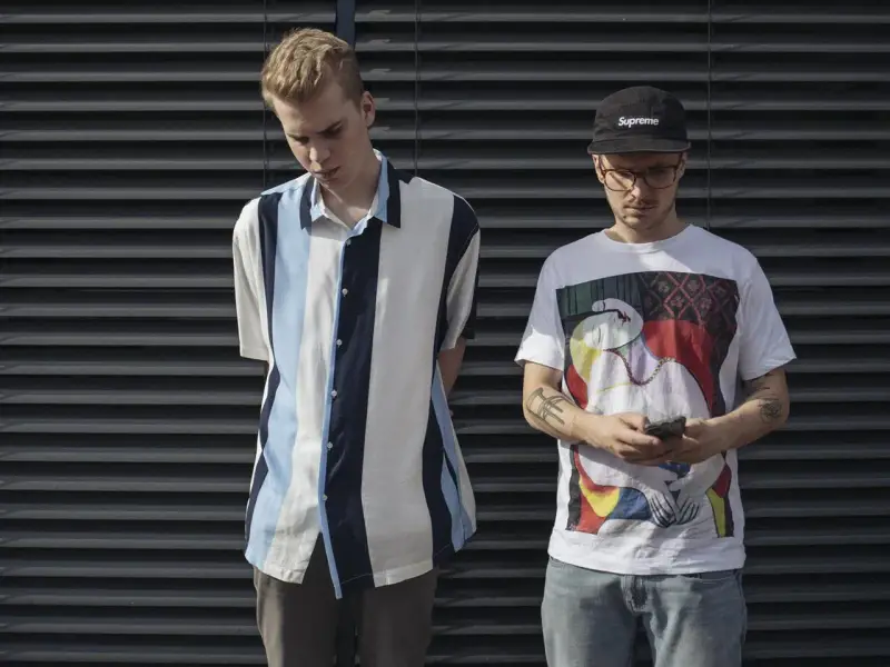 Dwóch młodych mężczyzn, jeden w koszuli w paski, drugi w koszulce z grafiką, jakby układanka Jana, stoi przed ciemną ścianą.